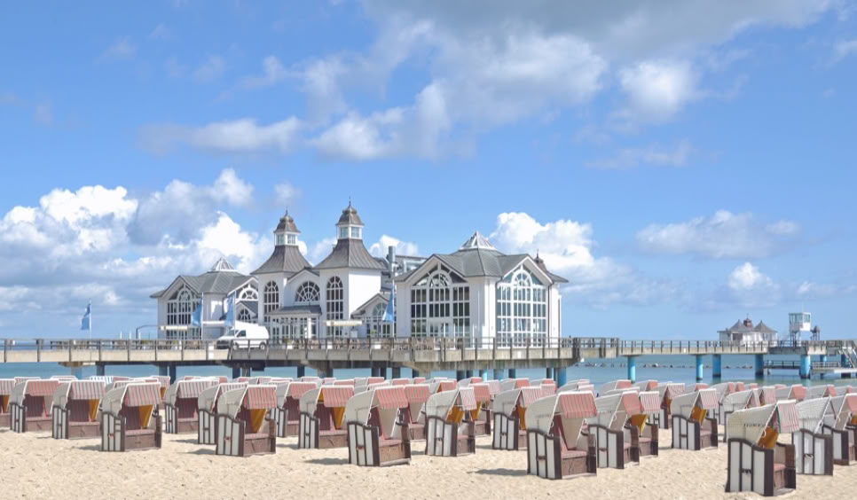 Strand mit Strandkörben im Vordergrund, dahinter ein Gebäude auf einer Seebrücke, blauer Himmel mit einigen Wolken.