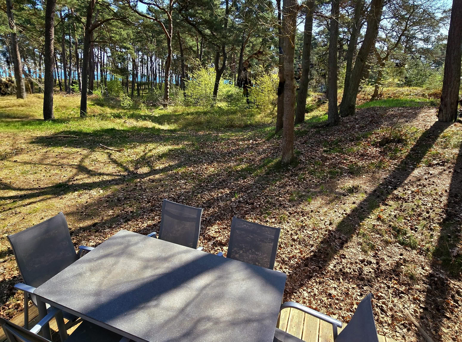 Terrasse mit einem Tisch und sechs rosa Stühlen, umgeben von einem weißen Geländer, neben einem hellblauen Gebäude. Im Hintergrund sind Bäume zu sehen.