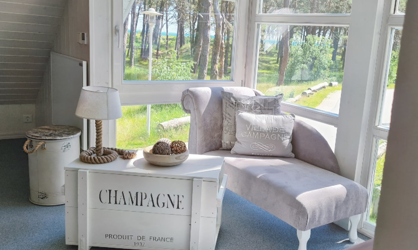 Gemütlicher Raum mit einem hellen Sessel, einem kleinen Tisch mit der Aufschrift "Champagne" und einem Fenster mit Blick auf Bäume.