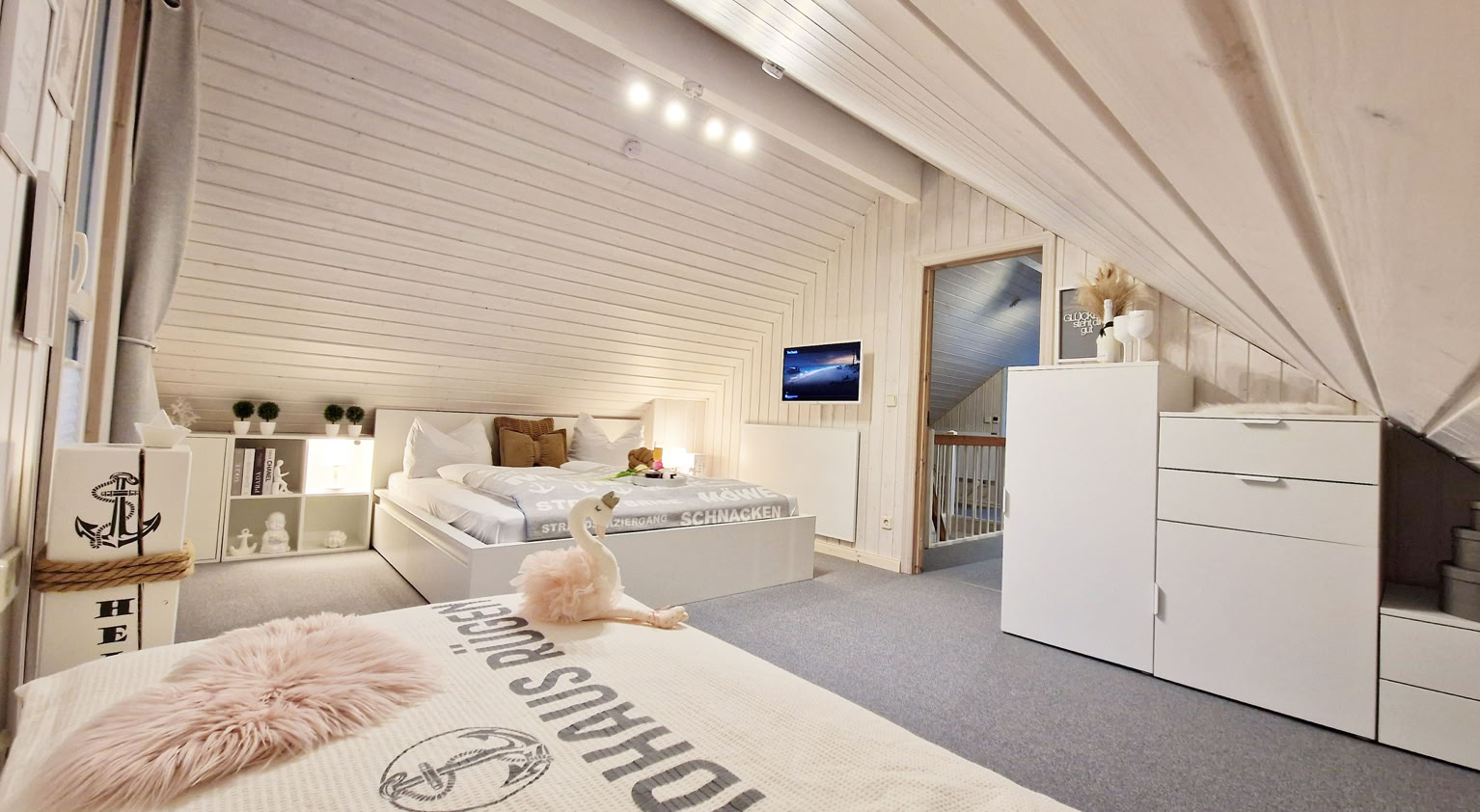 Ein Dachzimmer mit zwei Betten, weißen Möbeln, einem Fernseher an der Wand und schrägen Decken.