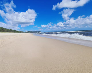 Strand mit hellem Sand, blauem Himmel und weißen Wolken, Wellen am Ufer.