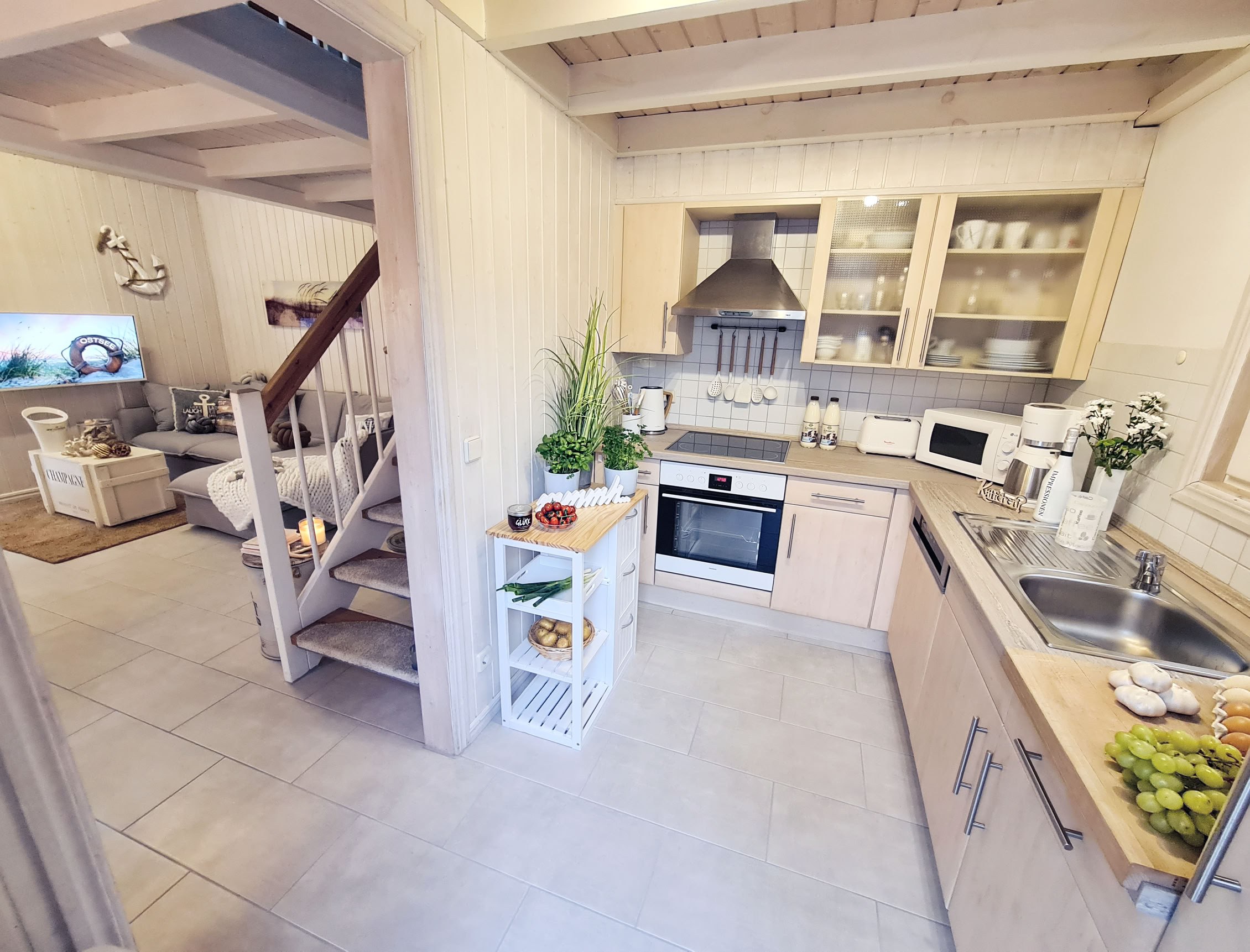 Kleine Küche mit Holzschränken, Herd, Mikrowelle und Spüle; daneben ein Wohnzimmer mit Sofa und Fernseher, Treppe führt nach oben.