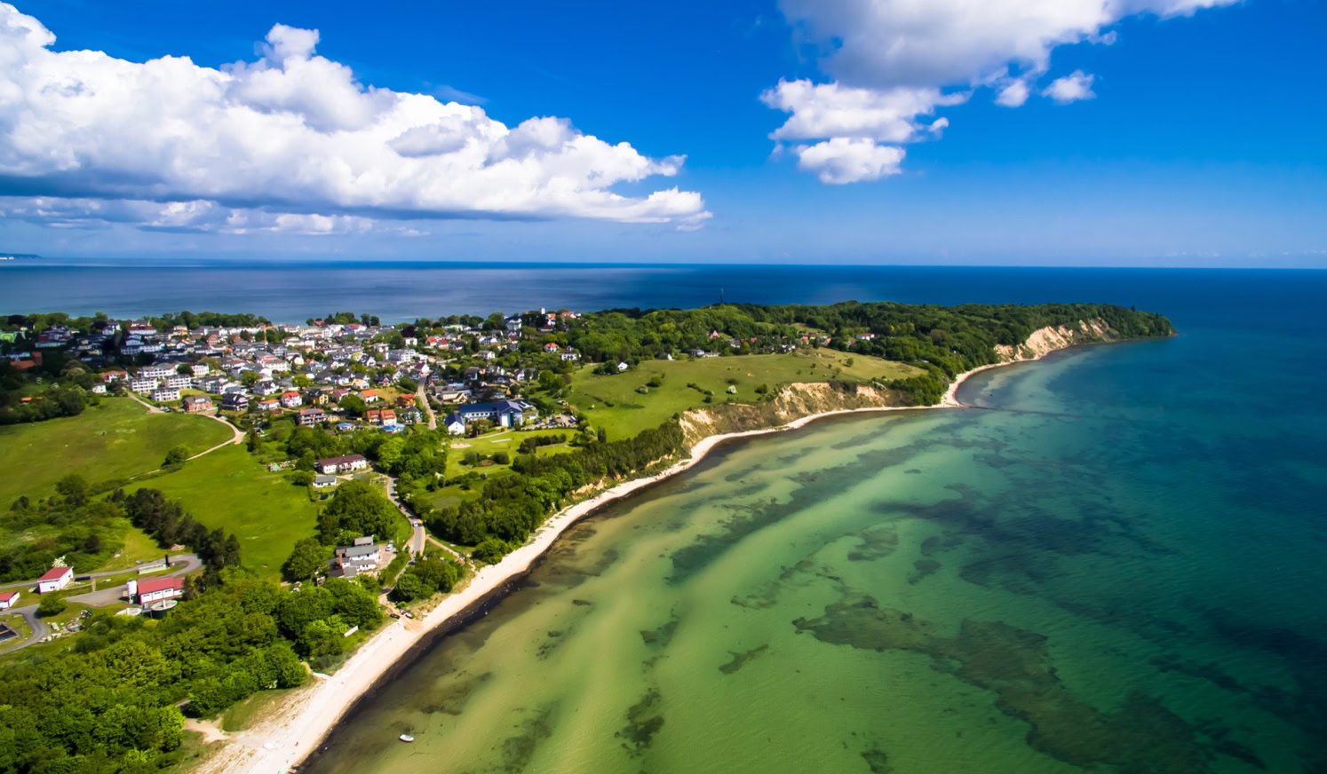 Luftaufnahme einer Küstenlandschaft mit grünem Wasser, Sandstränden, grünen Feldern und einer Siedlung im Hintergrund unter einem blauen Himmel mit weißen Wolken.