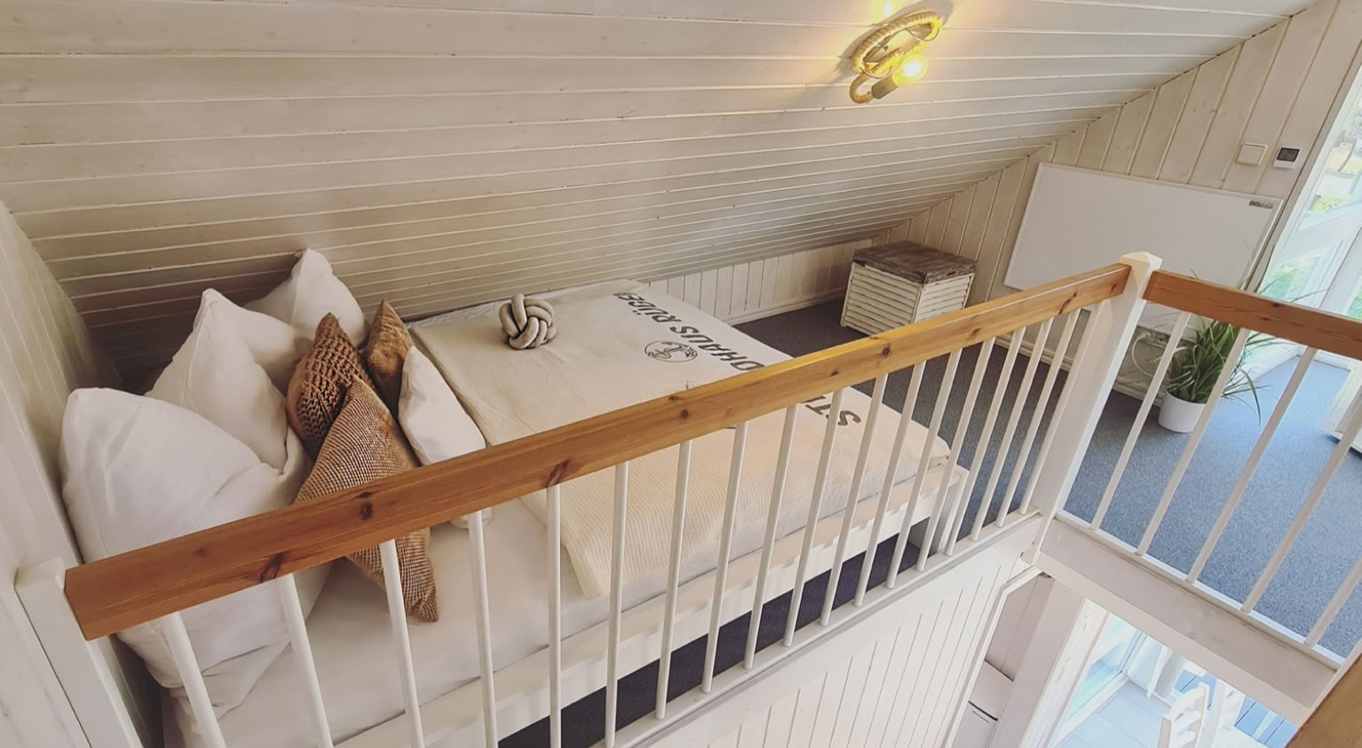 Ein kleines Schlafzimmer auf einer Empore mit einem Bett, weißen Kissen, einer Decke, einem Geländer und einer Lampe an der Decke. Darunter ist eine Treppe zu sehen.