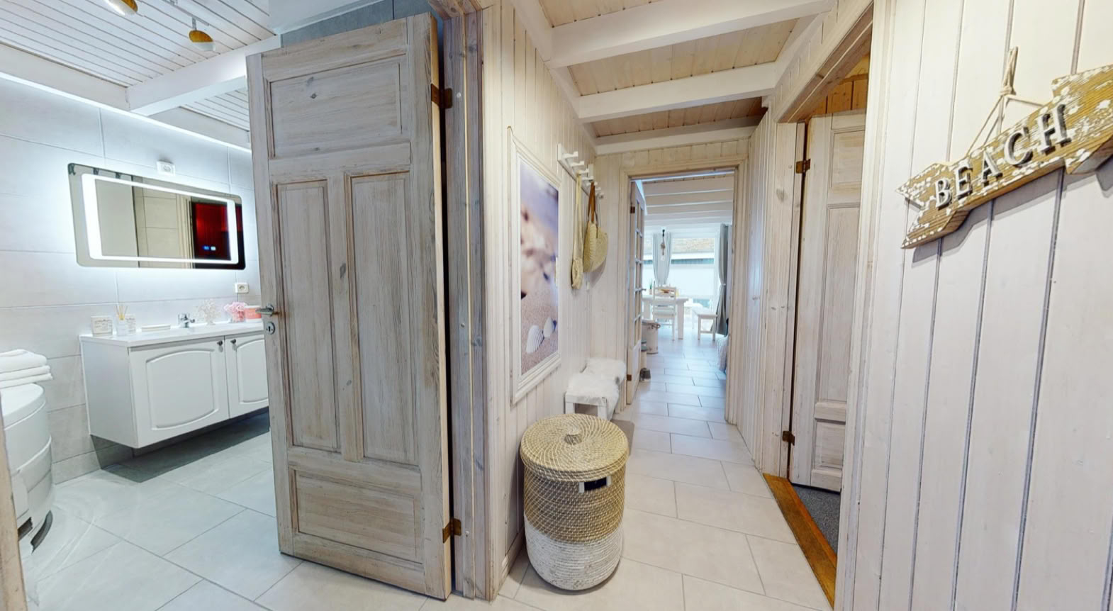 Ein heller Flur mit Holzwänden, einer offenen Tür zu einem Badezimmer links, einem Spiegel und einem Waschbecken. Rechts hängt ein Schild an der Wand.