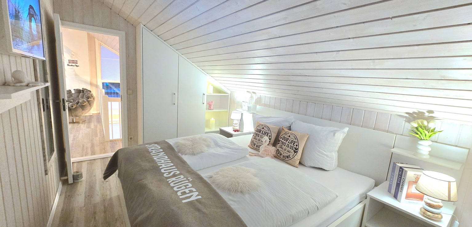 Gemütliches Schlafzimmer mit einem Doppelbett, blauen Bettdecken, mehreren Kissen, einem Nachttisch mit Lampe, einem Teppich und einem Fenster mit Blick nach draußen.