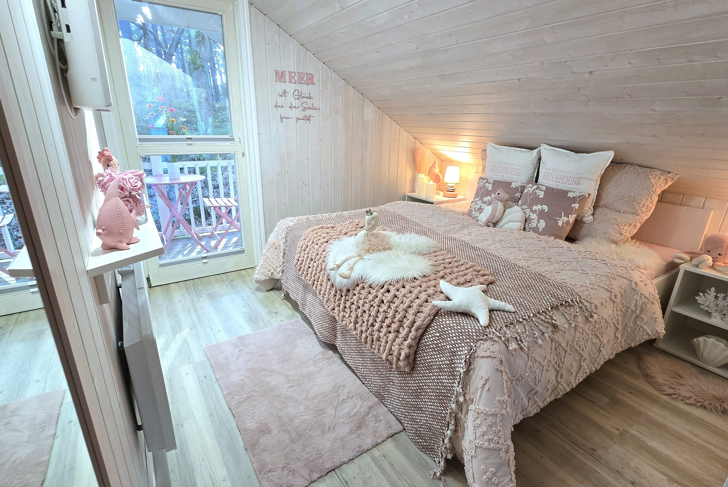Gemütliches Schlafzimmer mit Dachschräge, Doppelbett mit mehreren Kissen, Nachttischlampen, Fenster mit Blick nach draußen und dekorativen Elementen.