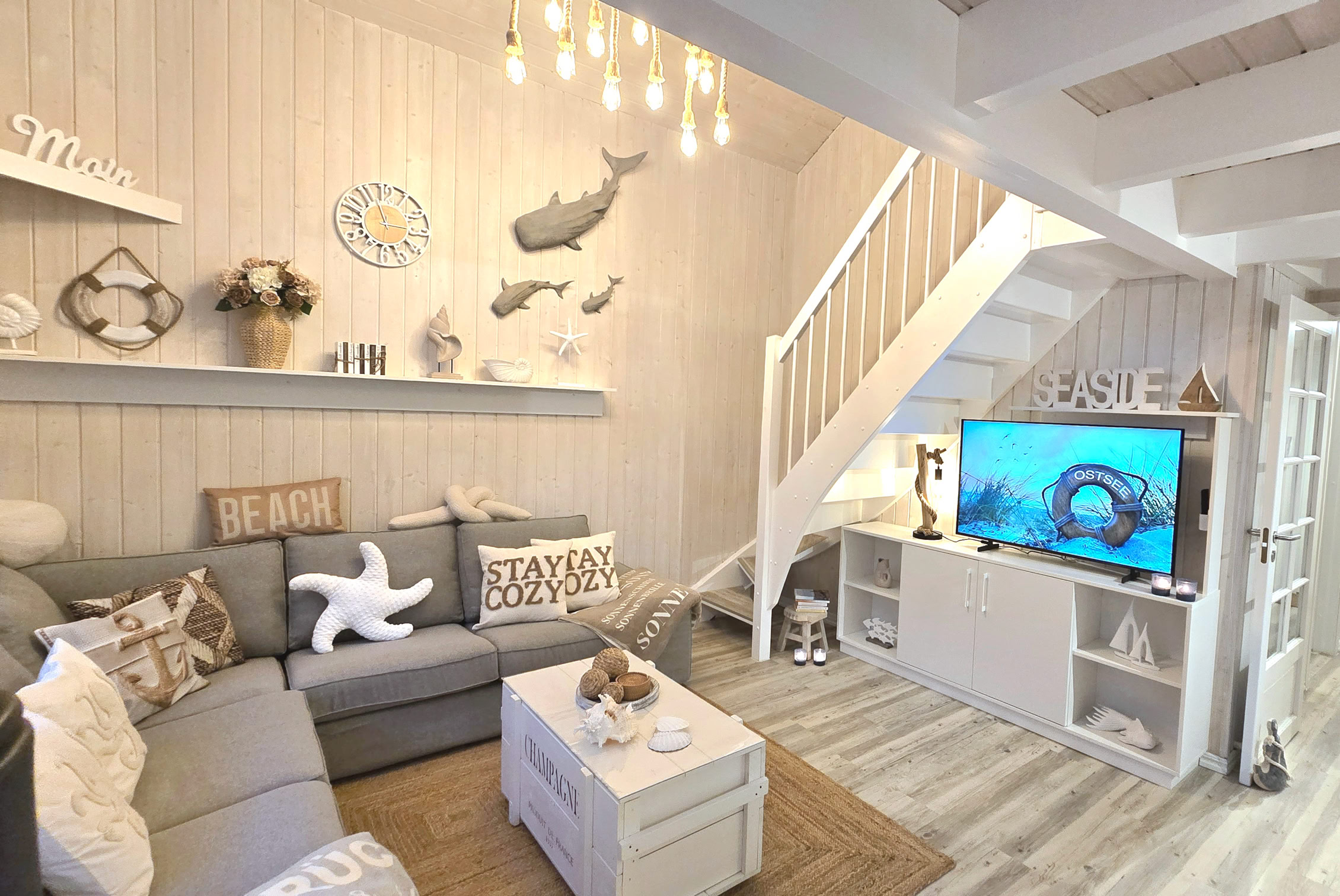 Wohnzimmer mit Kamin, Sofa, Couchtisch, dekorativen Kissen, Regalen an der Wand, Treppe und großen Fenstern.