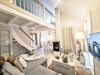 Helles Wohnzimmer mit grauem Sofa, Kamin, Treppe zu einer Galerie, Esstisch im Hintergrund und großem Fernseher.