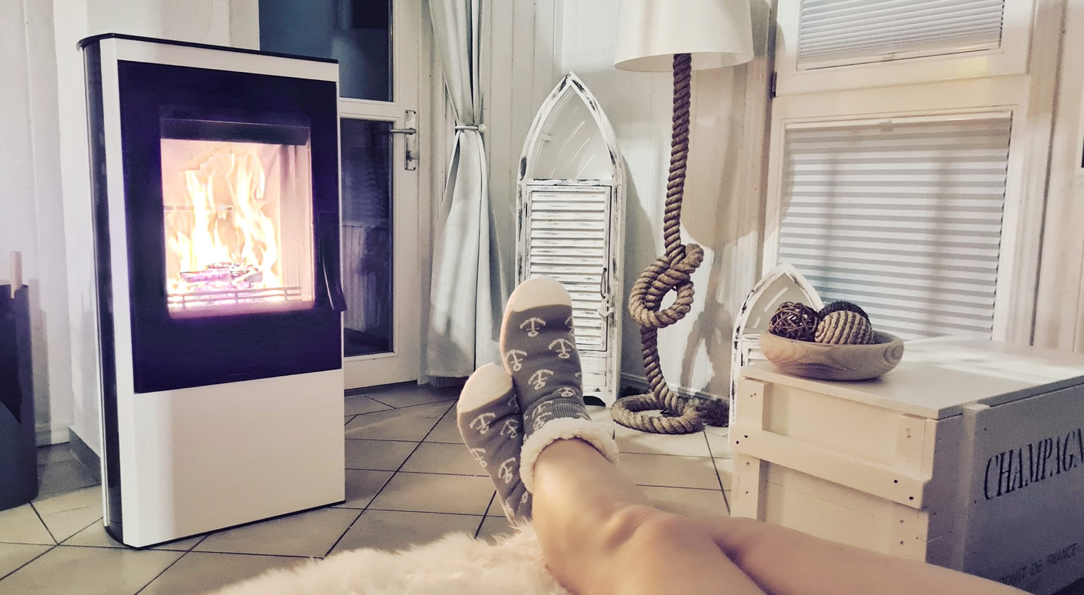 Beine einer Person auf einem Fellteppich vor einem Kaminofen, im Hintergrund ein Stuhl, eine Stehlampe und ein Fenster.